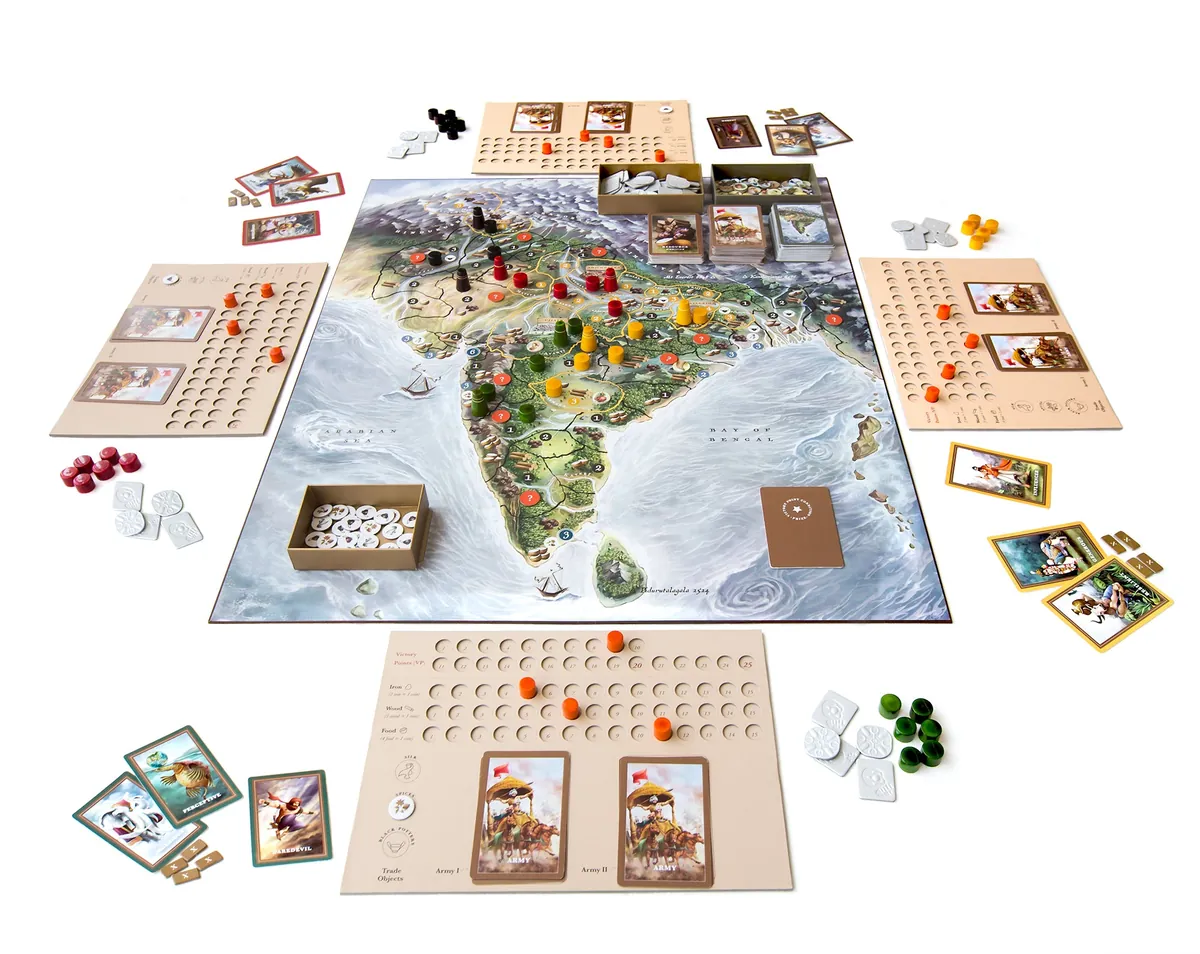 A four-player game set up on a white table: the large map of the Indian subcontinent in the middle, a player tracker board with wooden pegs on each side, card decks, a tray of coins, and wooden armies and forts in red, yellow, green and black