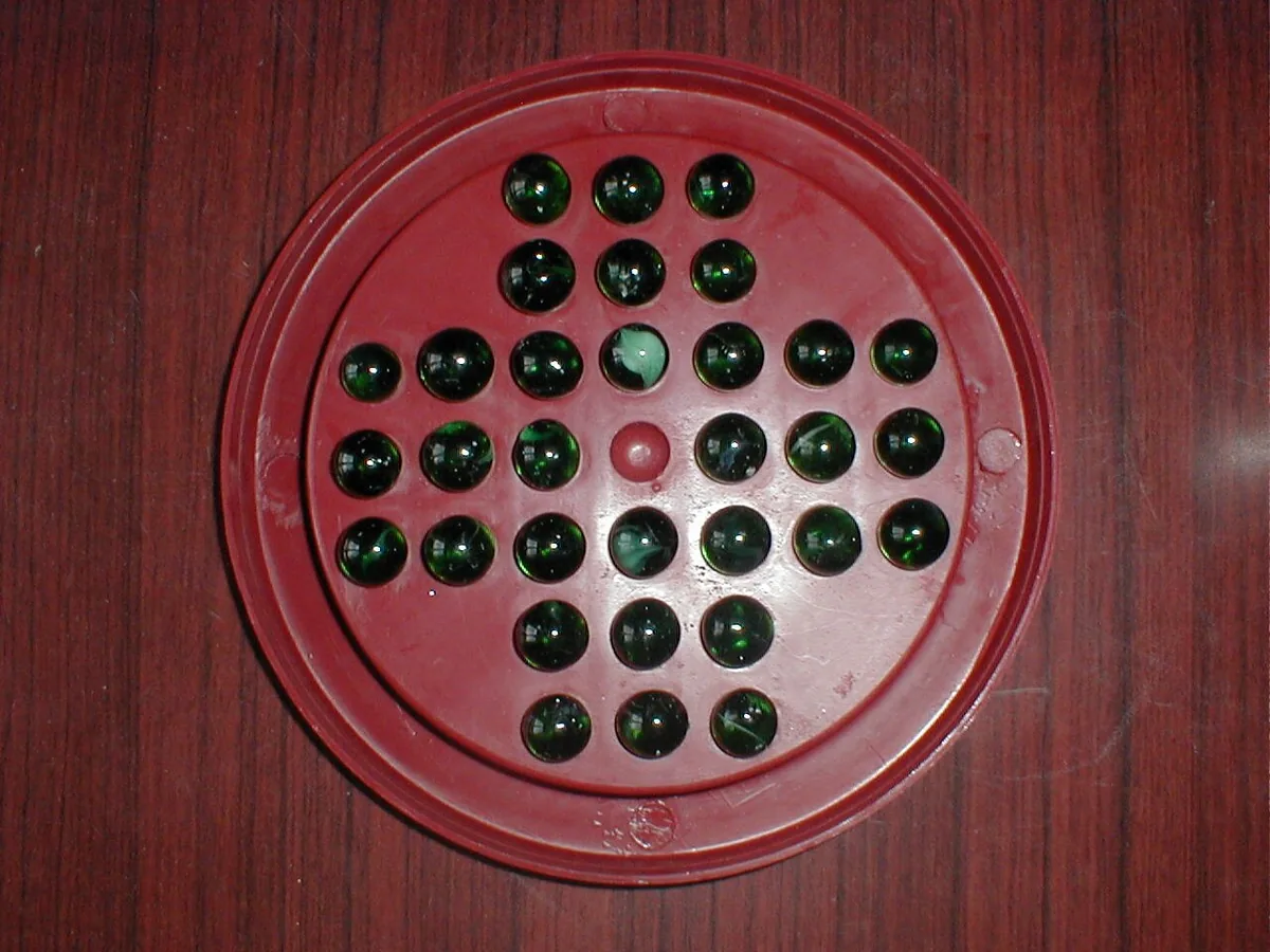 A red plastic Brainvita board with green glass marbles in 32 of its 33 holes and the centre hole empty