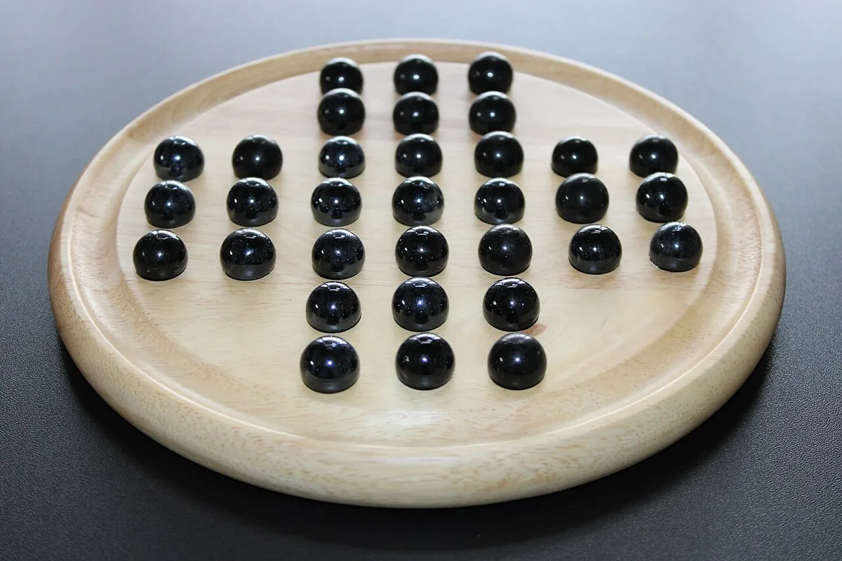 A round wooden peg solitaire board with black marbles, a few holes already empty