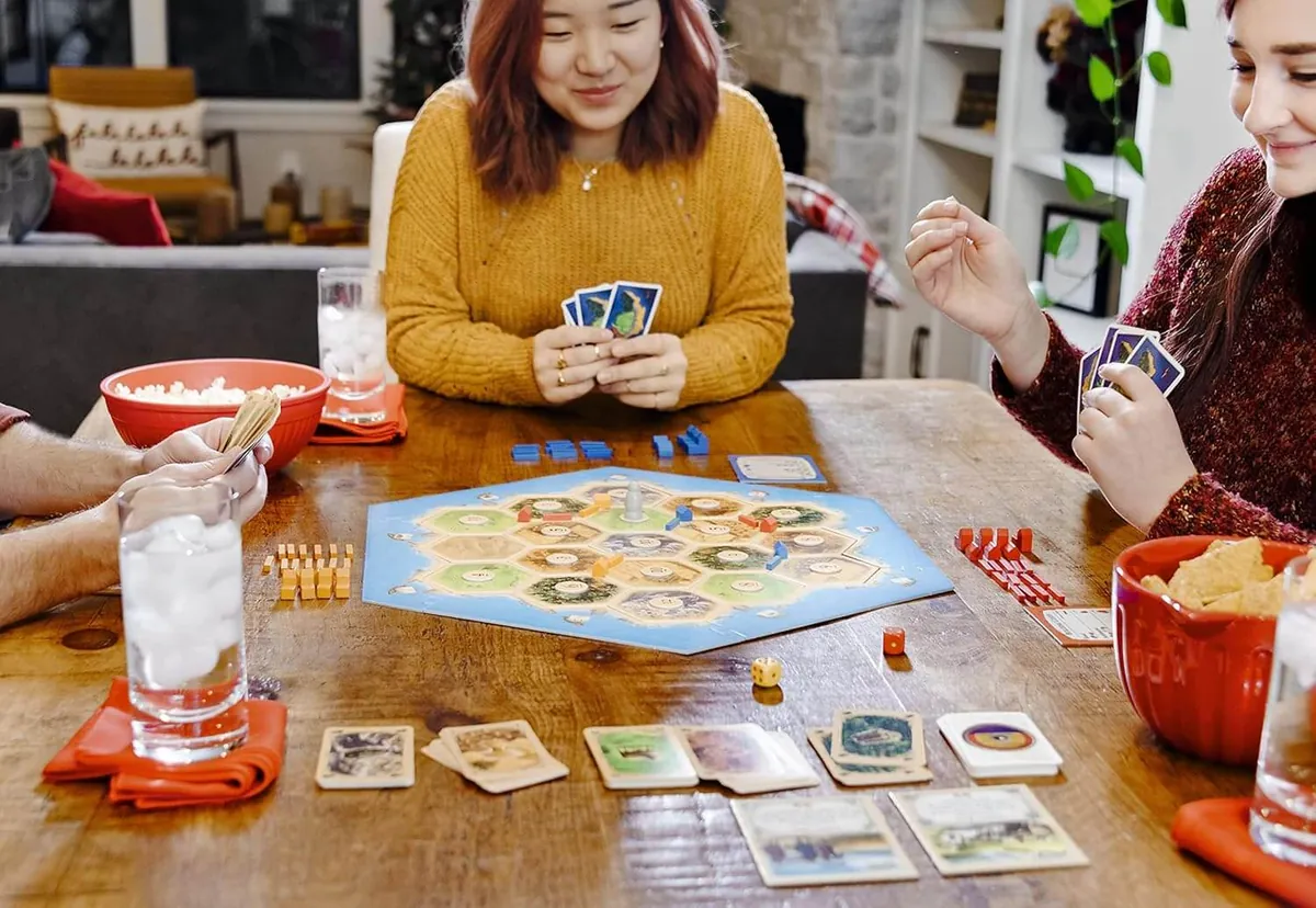 Friends playing Catan at a wooden table, one holding a fan of cards, with the board, cards and snacks in front of them