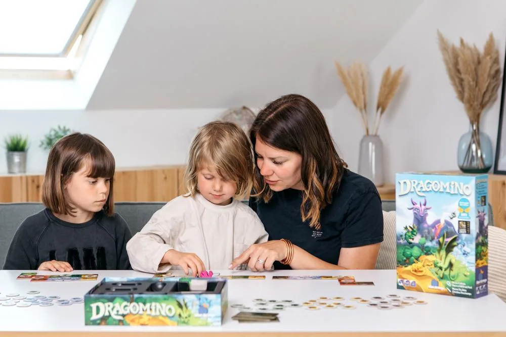 A woman and two children playing Dragomino, with the box on the table beside them