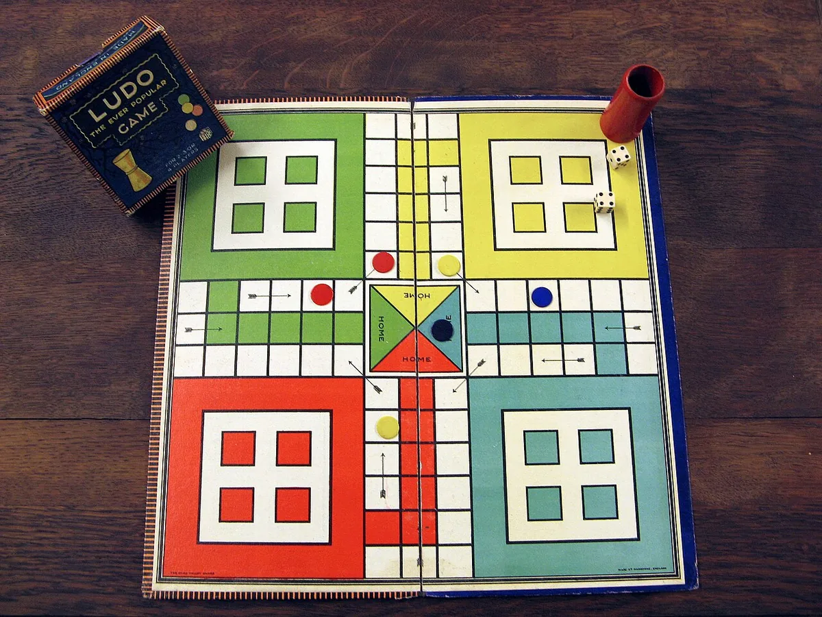 A printed Ludo board with coloured home squares and lanes, with counters and a die cup beside it