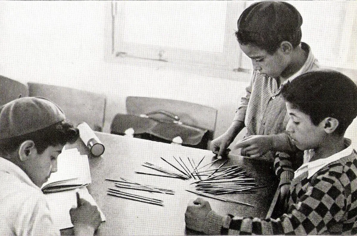 A black-and-white photo from the early 1950s of three boys playing pick-up sticks at a table