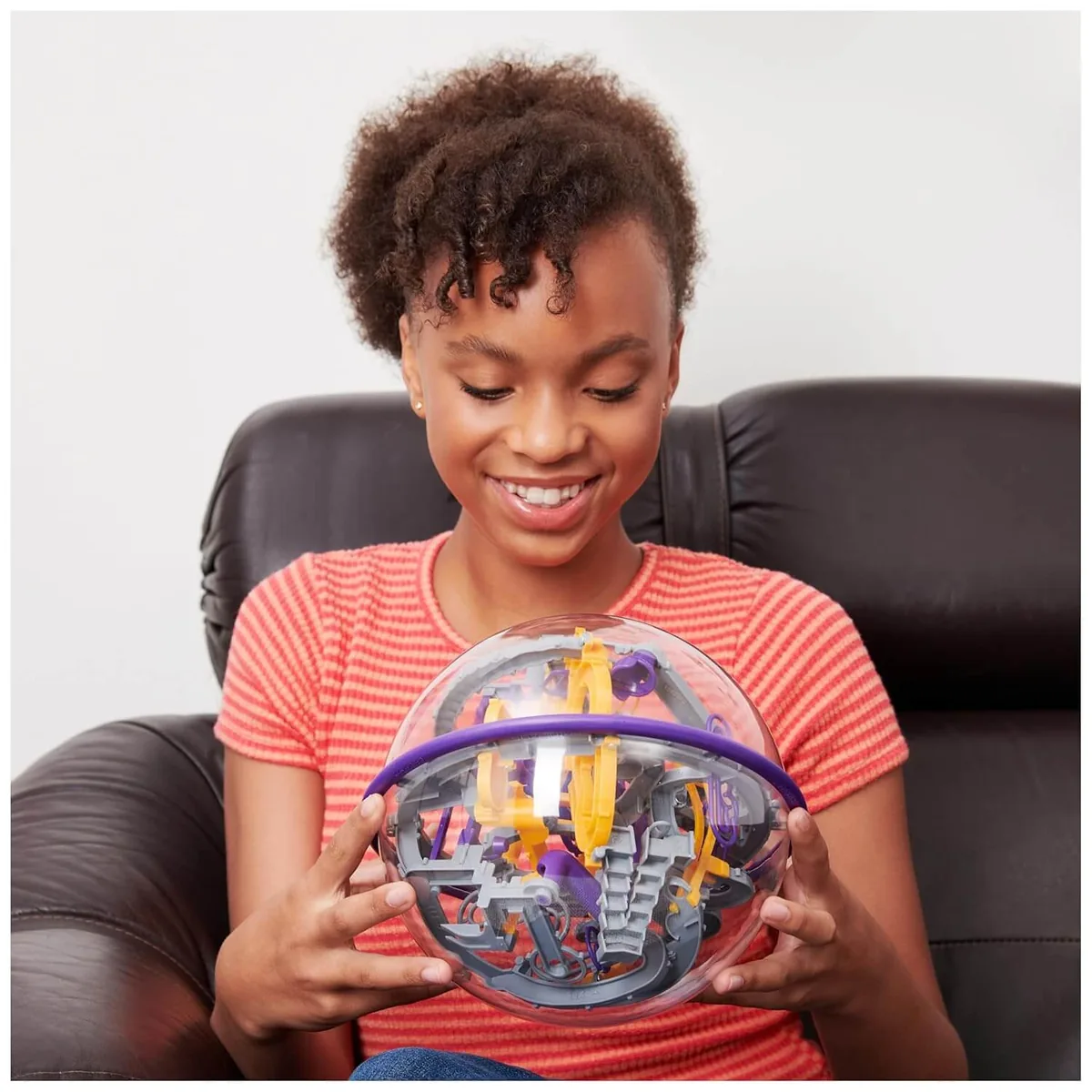 A smiling player in a striped top, sitting in an armchair and holding the Perplexus Epic sphere in both hands