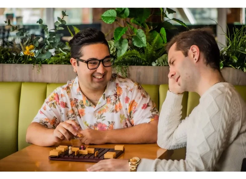 Two friends at a cafe table, one laughing and one thinking hard over a Quoridor board