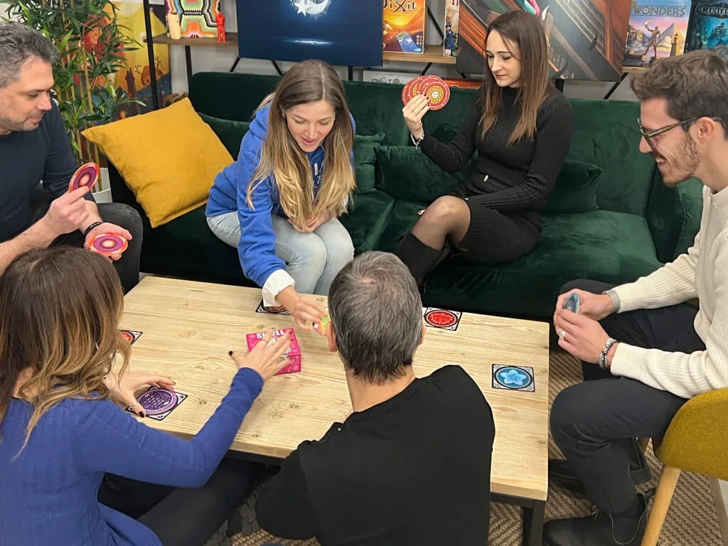 Six friends on sofas around a coffee table, holding Skull discs and making bids