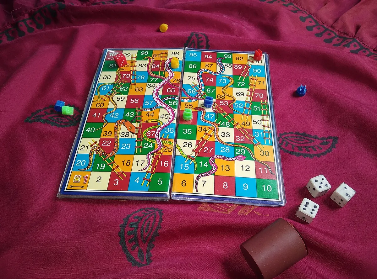 A Snakes and Ladders board numbered 1 to 100 on a red cloth, with dice and a dice cup