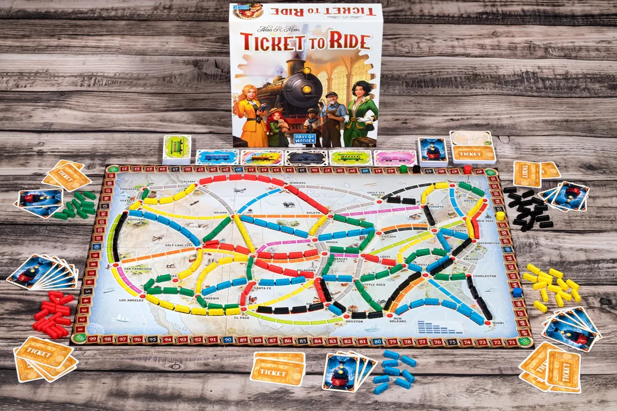 The board set up on a wooden table, with trains, card piles and tickets around it and the box behind