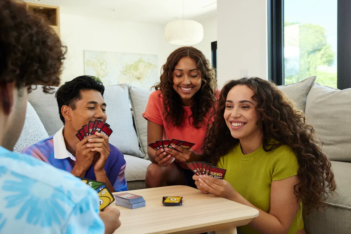 Three young adults laughing on a sofa around a coffee table, holding UNO No Mercy cards
